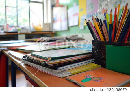 Colorful school books and stationery are stacked on a desk in a lively classroom with natural light streaming in from the windows 126438679