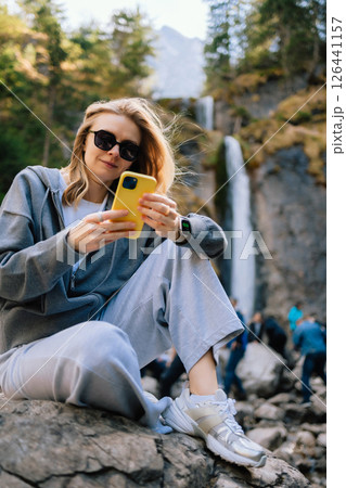 Blonde tourist wearing sunglasses and casual clothing is using mobile phone while sitting on rock near waterfall in mountain valley, enjoying nature and technology during hiking trip. Blonde hiker 126441157