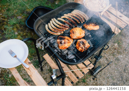 Sausages and chicken legs sizzling on hot barbecue, basking in sun, creating perfect setting for delightful outdoor meal in garden with family and friends. Grilled sausages and chicken, blazing coals Sausages and chicken legs sizzling on hot barbecue, basking in sun, creating perfect setting for delightful outdoor meal in garden with family and friends. Grilled sausages and chicken, blazing coals 126441158