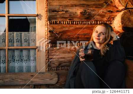 Young woman wrapped in cozy blanket, savoring warm drink while sitting by window of rustic wooden mountain cabin during stunning sunset, embracing tranquility and comfort. Girl sipping warm beverage Young woman wrapped in cozy blanket, savoring warm drink while sitting by window of rustic wooden mountain cabin during stunning sunset, embracing tranquility and comfort. Girl sipping warm beverage 126441171