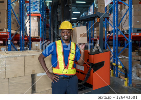 A man in safety gear hold a large cardboard box while standing in a well-organized warehouse. Shelve are lined with various product, indicating a busy working environment. 126443681