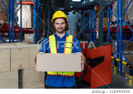A man in safety gear hold a large cardboard box while standing in a well-organized warehouse. Shelve are lined with various product, indicating a busy working environment. A man in safety gear hold a large cardboard box while standing in a well-organized warehouse. Shelve are lined with various product, indicating a busy working environment. 126444350