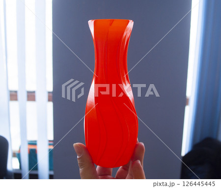 Person holding red vase model, printed on 3D printer, against blue wall. Three-dimensional model printed on 3D printer from molten plastic of red color. Concept 3D Printing. FDM 3D Printing technology Person holding red vase model, printed on 3D printer, against blue wall. Three-dimensional model printed on 3D printer from molten plastic of red color. Concept 3D Printing. FDM 3D Printing technology 126445445