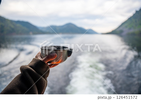 Hand holding hot drink in metal mug with view of Teletskoye lake in Altai Russia. Cozy moment in wild mountain nature during outdoor travel 126451572