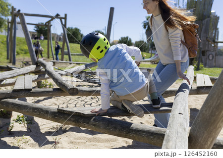 Child wearing helmet crawling on wooden playground structure with mother supervising 126452160