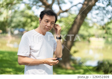 Wellness and Mindfulness. Senior man adjusting earbuds while enjoying nature in the park. Wellness and Mindfulness. Senior man adjusting earbuds while enjoying nature in the park. 126452809