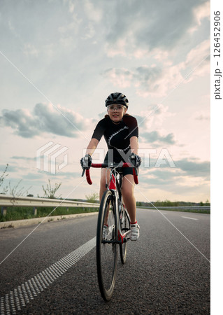 Smiling female cyclist, dressed in athletic gear and safety helmet, rides road bike confidently along highway curve in evening light, outdoor. Smiling female cyclist, dressed in athletic gear and safety helmet, rides road bike confidently along highway curve in evening light, outdoor. 126452906