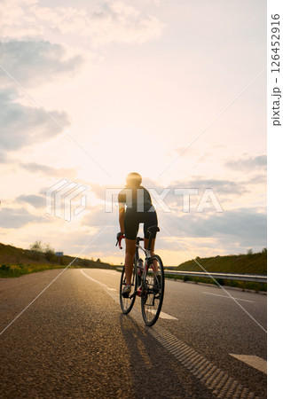 Sporty cyclist riding along highway in evening light, confident posture and silhouette glowing under dramatic sky full of soft clouds Sporty cyclist riding along highway in evening light, confident posture and silhouette glowing under dramatic sky full of soft clouds 126452916