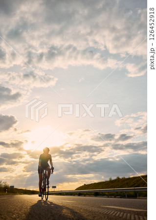 Woman cycling directly into sun rays in scenic open road at sunset, framed by dramatic sky and strong light burst. Marathon. 126452918