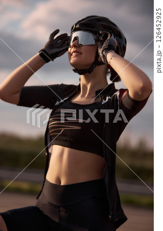 Smiling female athlete adjusting reflective helmet and sunglasses while enjoying warm sunset light after bike ride, intense training outdoor. 126452925