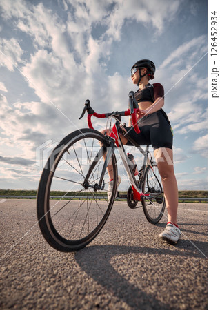 Low angle view photo of woman standing next to racing bike, one foot forward, glancing into distance, sky filled with soft clouds in background. Low angle view photo of woman standing next to racing bike, one foot forward, glancing into distance, sky filled with soft clouds in background. 126452934