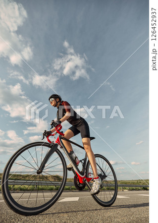 Low angle view photo of female cyclist leaning into motion on sunny highway, aerodynamic pose highlight action. 126452937