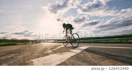 Low angle view photo of cyclist riding fast toward horizon under bright sun, with curved road and glowing sky enhancing dynamic sense of speed. 126452940