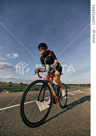 Low angle view photo of female cyclist riding on curvy road, hands on lower handlebars, showing aggressive racing posture and focus in eyes. 126452943