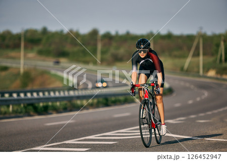 Dynamic photo of determined female cyclist approaching uphill curve on open road, pushing pedals in intense focus, wearing helmet and tight jersey. Dynamic photo of determined female cyclist approaching uphill curve on open road, pushing pedals in intense focus, wearing helmet and tight jersey. 126452947