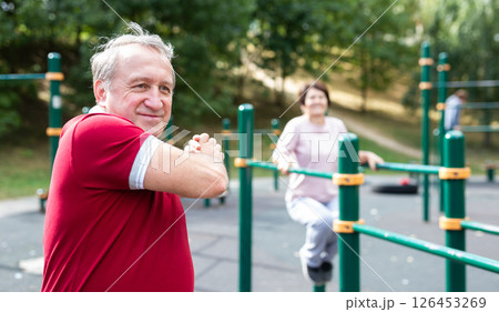 Staying after retirement. Happy joyful mature retired sportsman wearing sportswear doing side stretching exercises with arm over his head, exercising outside in city park 126453269