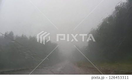 Heavy rain blurs view through car windshield, obscuring road ahead and creating misty atmosphere. Water droplets create textured effect on window, making visibility challenging Heavy rain blurs view through car windshield, obscuring road ahead and creating misty atmosphere. Water droplets create textured effect on window, making visibility challenging 126453311
