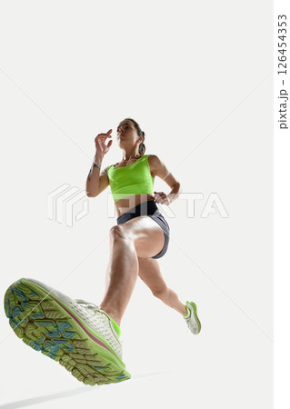 Low angle shot of female runner in bright green top and black shorts, stepping forward with powerful stride. Low angle shot of female runner in bright green top and black shorts, stepping forward with powerful stride. 126454353