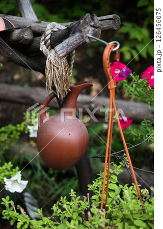 A rustic clay jug hangs from a wooden structure with a rope, surrounded by green plants and vibrant flowers. Concept of rural charm and nature 126456075