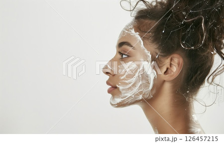 Young woman applying face mask while looking to the side in a bright indoor setting during a self-care routine 126457215