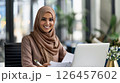 Smiling woman in a hijab working on a laptop in a modern office environment during daytime 126457602