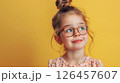 Young girl with glasses smiling against a bright yellow background, showcasing her playful spirit and innocence 126457607