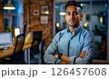 Young professional in a modern office setting confidently poses with arms crossed in a tech-driven workspace during the evening 126457608