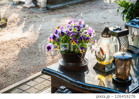 Vibrant pansies in pot on marble headstone amidst sunlit cemetery with ornate lanterns and intricate metal decorations reflect solemn memory 126458276