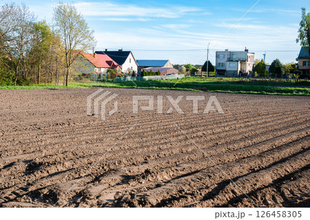 Expansive tilled field stretching towards distant houses under clear blue sky signaling spring's arrival in rural countryside 126458305