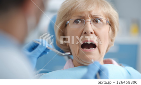 Mature patient showing anxiety, gripping dental chair armrests, experiencing dental checkup stress with medical tools nearby 126458800