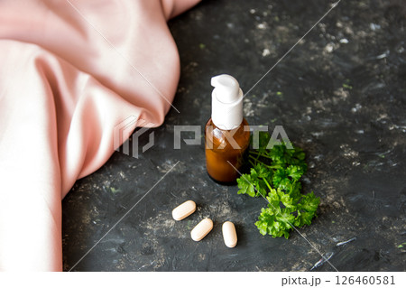 Brown glass bottle with essential oil or creme, vitaminen and healing green herbs on the rough texture background with pink silk fabric. 126460581