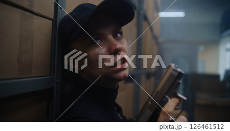 Close Up of Female Secret Agent Hiding Behind Cover in Warehouse, Holding a Pistol, Talking to Killer 126461512