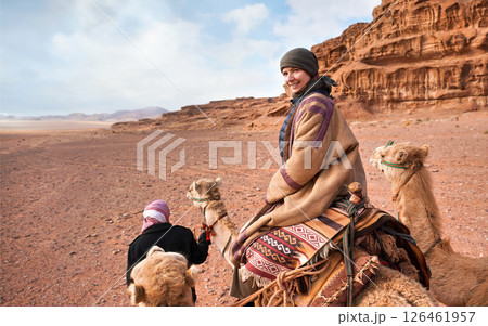 Young woman riding camel in Wadi Rum desert, looking back over her shoulder, smiling. It's quite cold so she is wearing traditional Bedouin coat - bisht - and head scarf, mountains far background 126461957