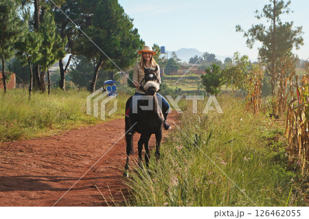 Young woman in shirt and straw hat, riding black horse in the park, blurred background with houses and trees Young woman in shirt and straw hat, riding black horse in the park, blurred background with houses and trees 126462055