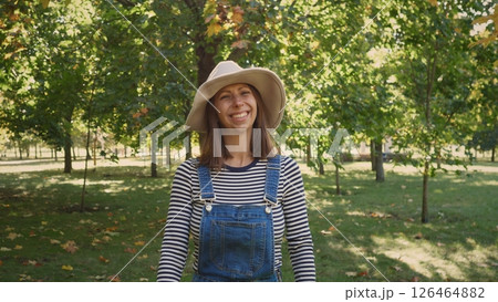 A woman wearing a striped long-sleeve shirt and denim overalls smiles in a vibrant park filled with green trees and autumn-colored leaves on a sunny day. 126464882