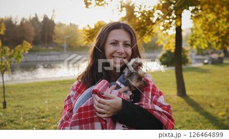 Smiling woman holds a small dog wrapped in a blanket as they share a warm moment in a park surrounded by colorful autumn leaves and a calm lake. Smiling woman holds a small dog wrapped in a blanket as they share a warm moment in a park surrounded by colorful autumn leaves and a calm lake. 126464899