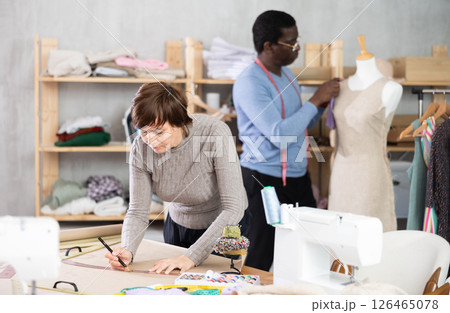 Female dressmaker sketching sewing pattern while colleague adjusting dress on mannequin Female dressmaker sketching sewing pattern while colleague adjusting dress on mannequin 126465078