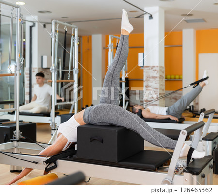 Girl works out in gym and performs set of exercises to stretch leg muscles with help of reformer Girl works out in gym and performs set of exercises to stretch leg muscles with help of reformer 126465362