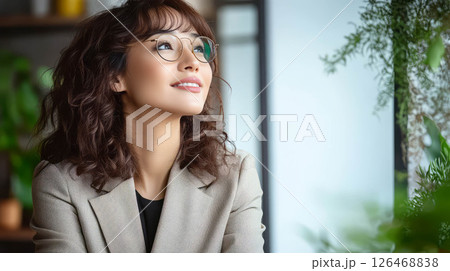 A young woman with glasses smiles thoughtfully in a cozy, plant-filled cafe setting. 126468838