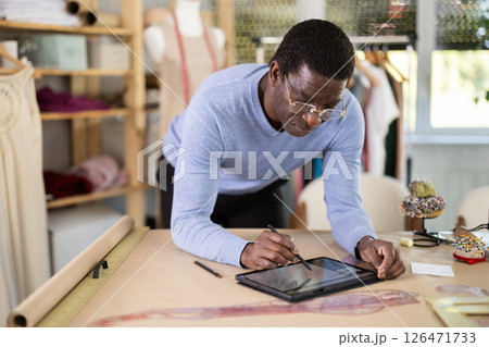 Male tailor draws sketch of future clothing on an electronic tablet. Working with modern gadgets in sewing workshop 126471733