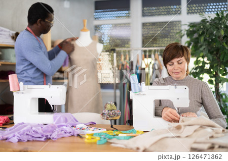 Woman tailor sews clothes, African male companion work in background with mannequin Woman tailor sews clothes, African male companion work in background with mannequin 126471862