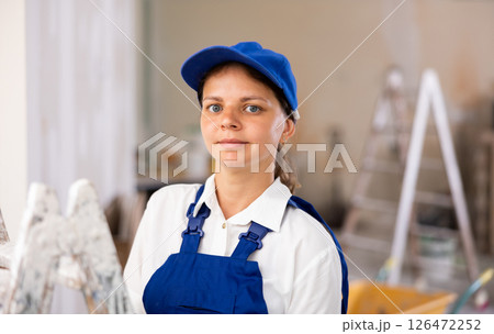 Portrait of contented young female builder in blue overalls during the repair of cottage 126472252