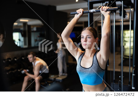 Slender athletic girl work out muscles of arms and shoulders on simulator in gym of fitness club 126472472