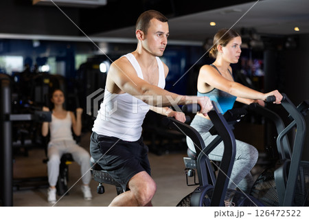 Sportive young couple doing air bike exercises in well-equipped gym Sportive young couple doing air bike exercises in well-equipped gym 126472522