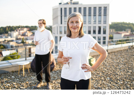 Happy senior woman with gray hair holding water bottle on rooftop, enjoying outdoors. Older man in background. Image conveys wellness, relaxation, activity, outdoor leisure, urban environment 126473242