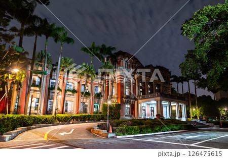Historic red-brick building of the National Taiwan University Hospital illuminated at night in Taipei, featuring neoclassical architecture framed by palm trees and a moody clouded sky. 126475615