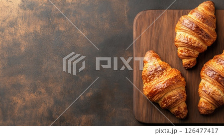 Golden baked croissants on rustic wooden cutting board against dark textured background, fresh pastry concept with copy space 126477417