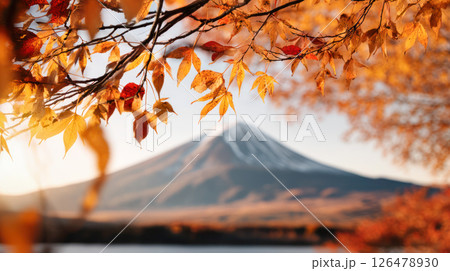 Autumn leaves in vibrant hues frame majestic snow-capped Mount Fuji peak, creating a stunning natural landscape scene with warm sunlight illuminating the tranquil lake below 126478930