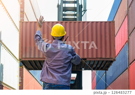 Dock supervisor in yellow hard hat and uniform directing container loading with walkie talkie and hand gesture at shipping container yard. Dock supervisor in yellow hard hat and uniform directing container loading with walkie talkie and hand gesture at shipping container yard. 126483277