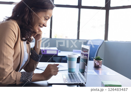 Smiling woman using laptop at office desk with coffee cup nearby Smiling woman using laptop at office desk with coffee cup nearby 126483487